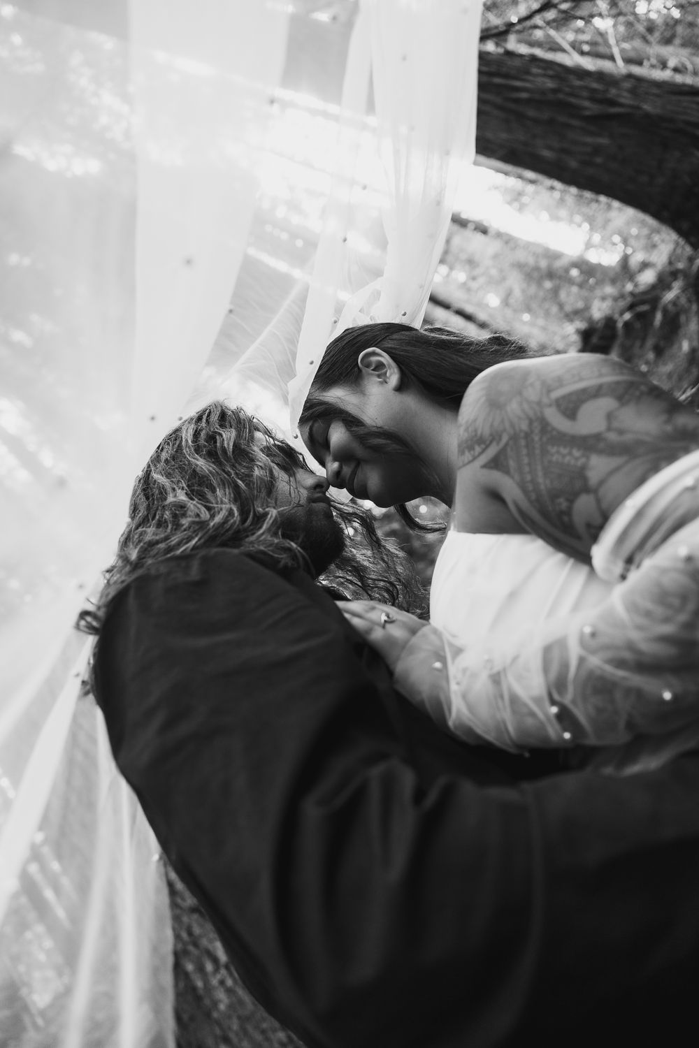 Bride and groom share an intimate moment, nearly kissing, surrounded by a flowing white veil outdoors.