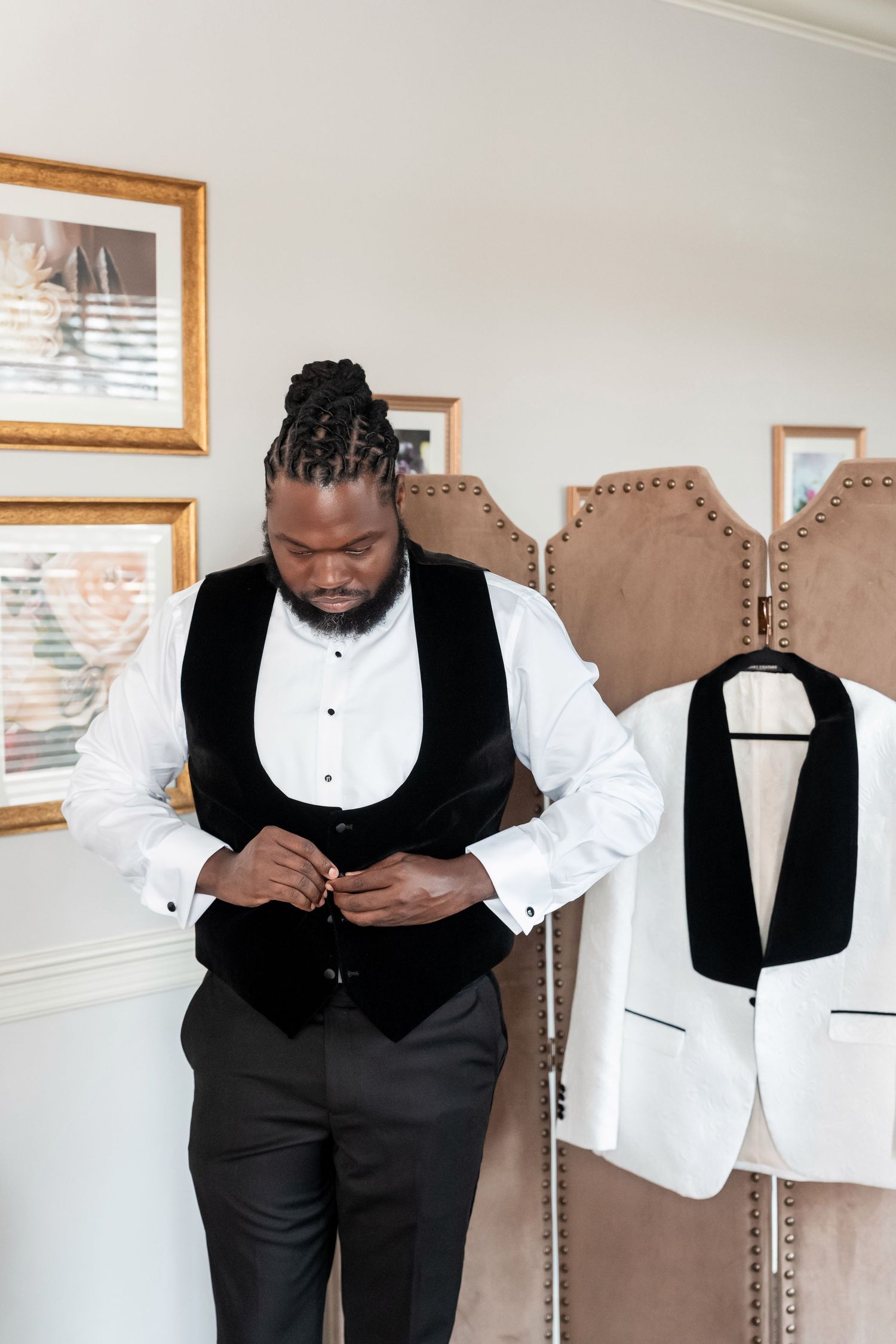 A groom in a white shirt and black velvet vest buttons up while a white tuxedo jacket hangs nearby. Deerfield Golf Club Newark De