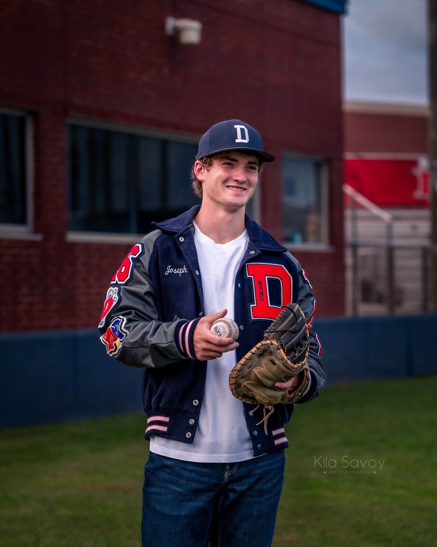 Young baseball player in a varsity jacket and cap holds a glove and ball, smiling outdoors near a brick building.