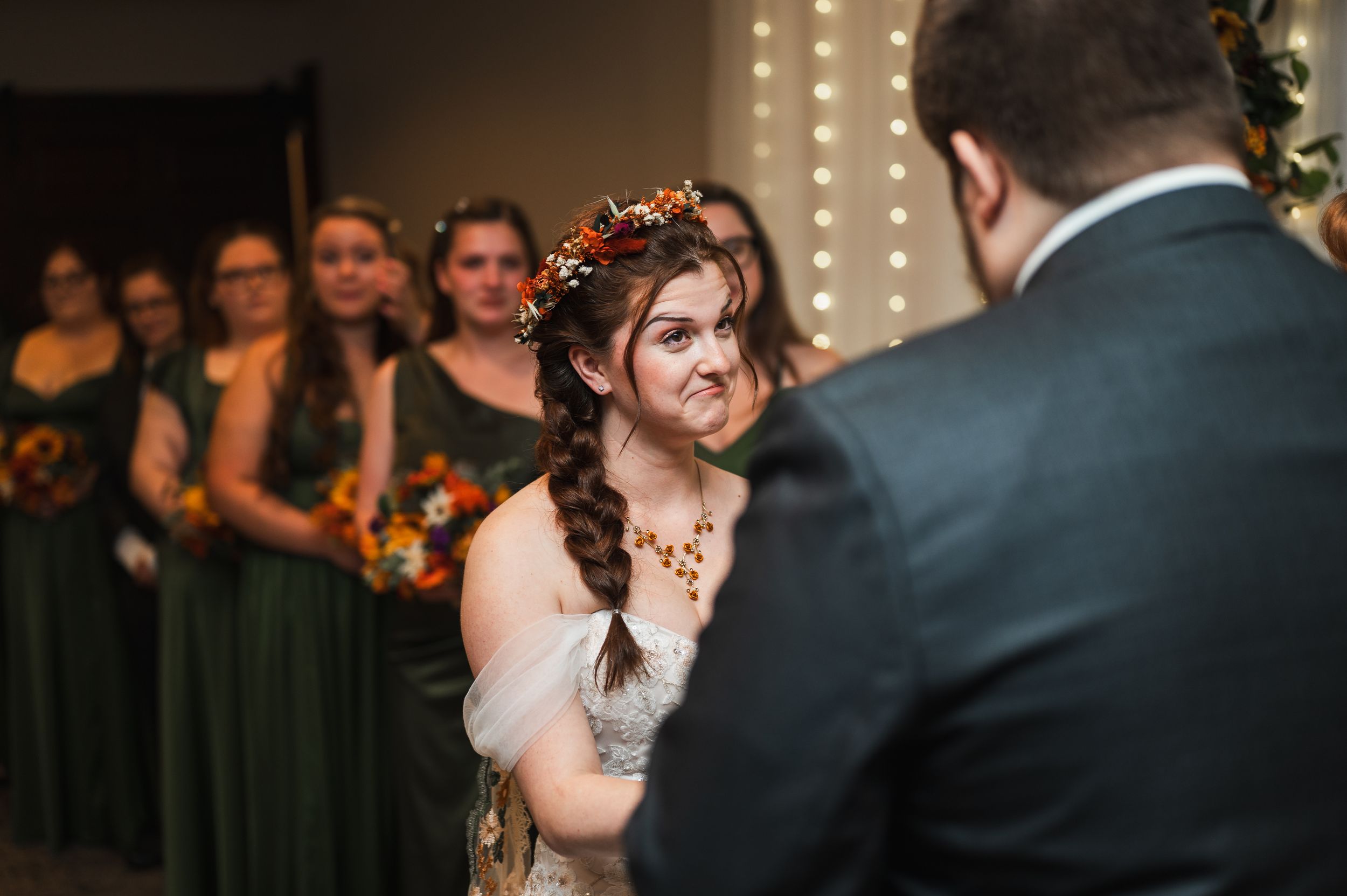 Bride in floral crown smiles at groom during wedding ceremony with bridesmaids in green dresses in background.