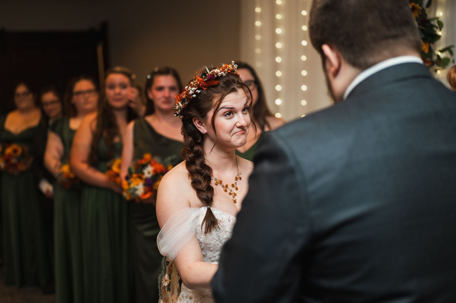 Bride in floral crown smiles at groom during wedding ceremony with bridesmaids in green dresses in background.