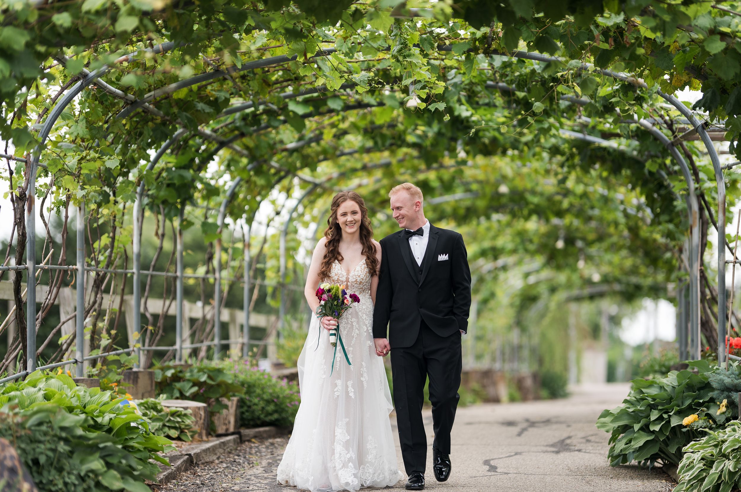 Bride and groom walking hand in hand under a lush green garden archway on their wedding day.