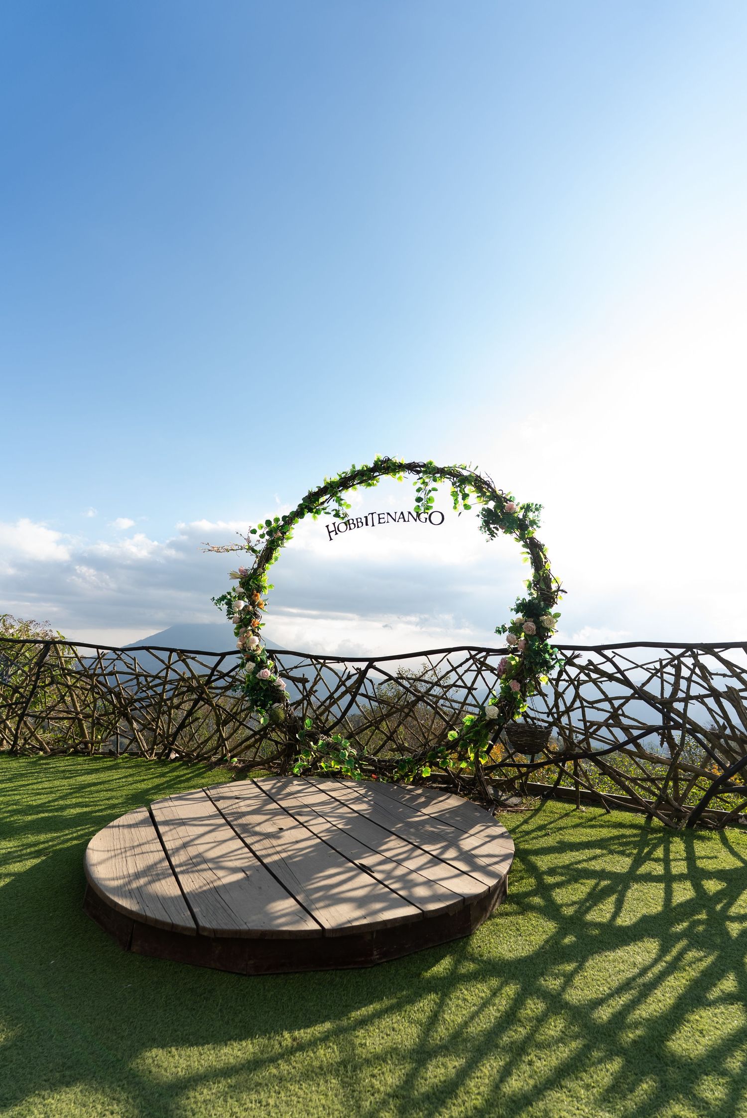 A floral wedding arch with a circular wooden platform overlooks a scenic mountain landscape on a sunny day.