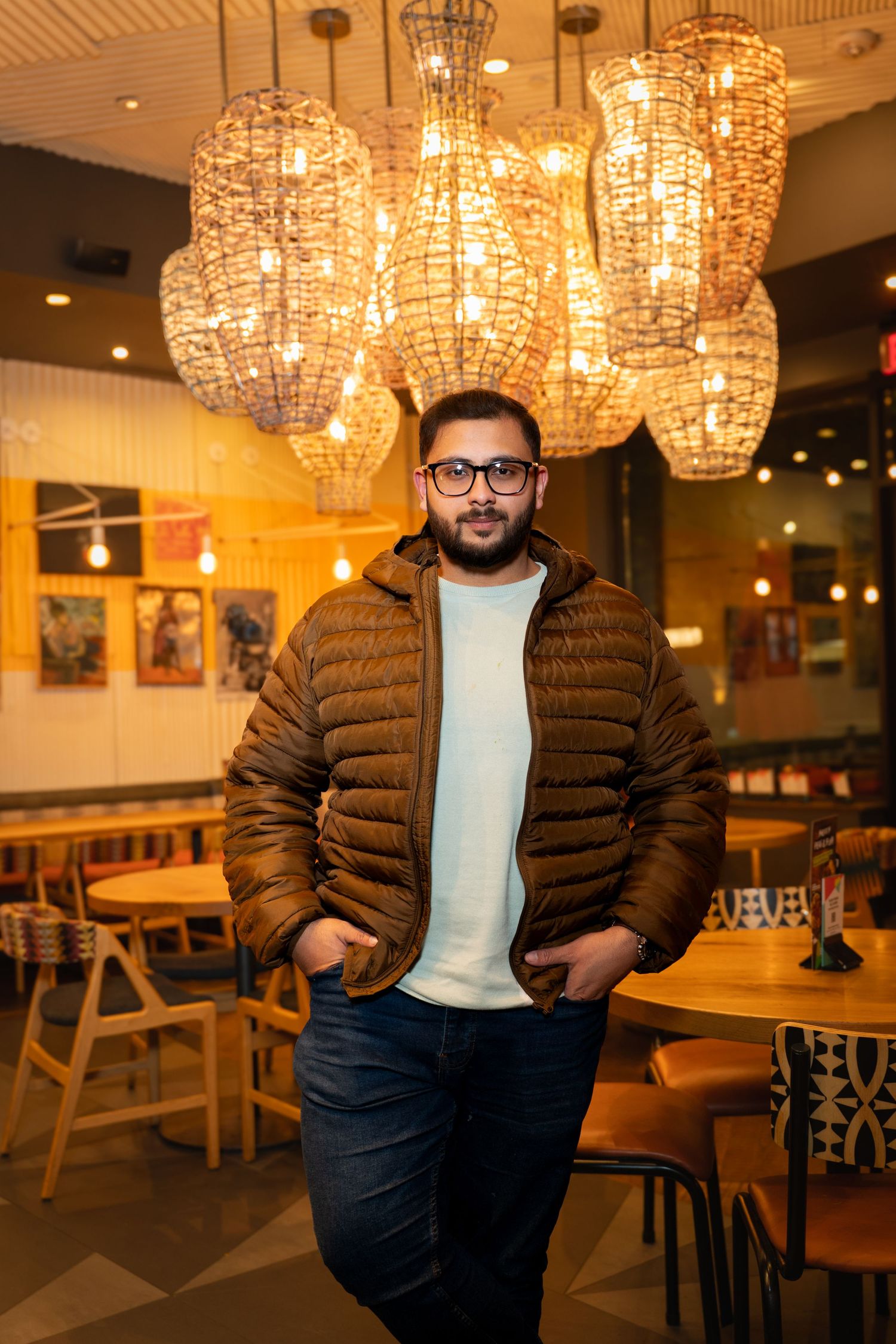 Person in brown puffer jacket and glasses sits in warmly lit restaurant with crystal chandeliers overhead.