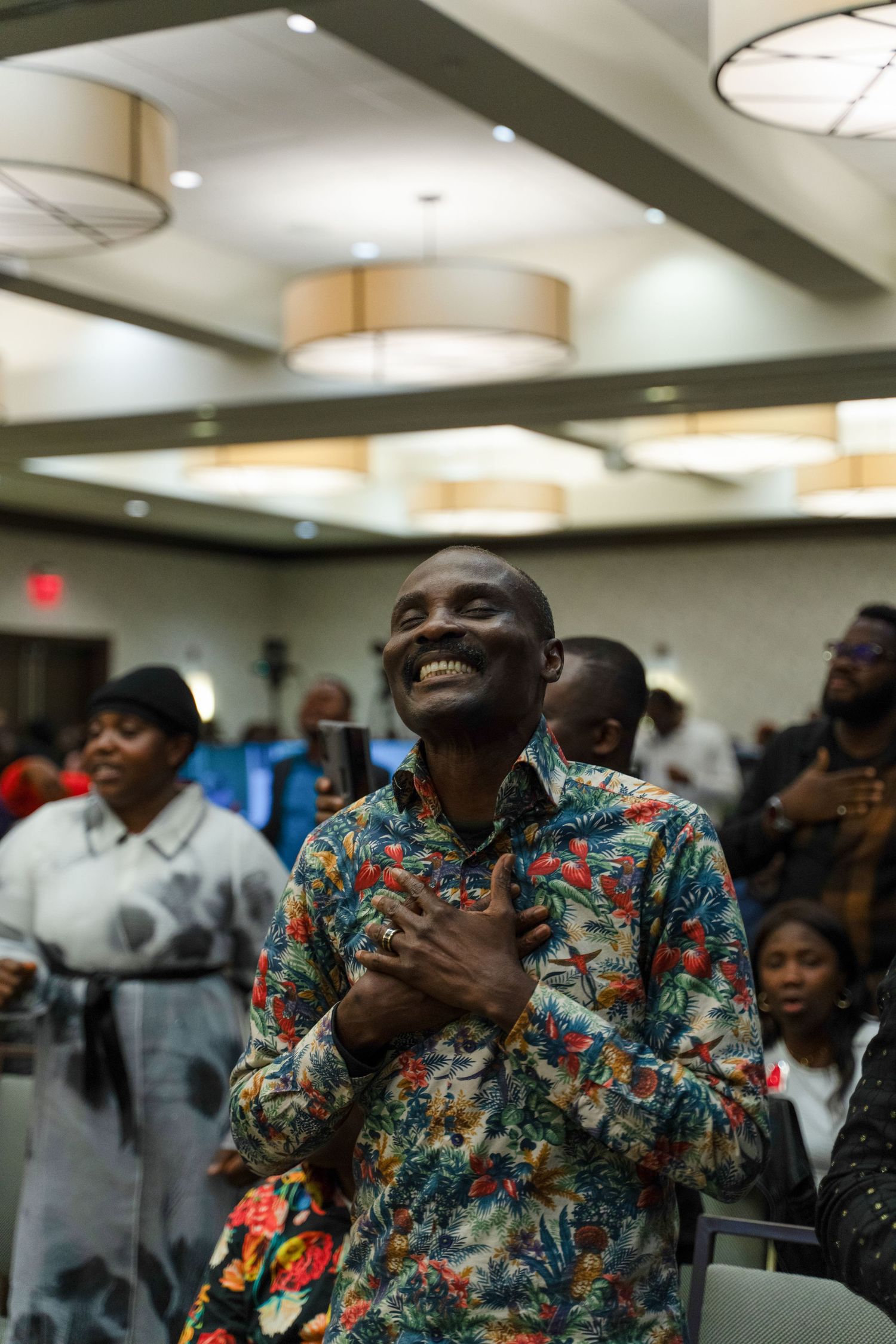 A person in a colorful patterned shirt smiles with joy during an emotional moment at an indoor gathering.