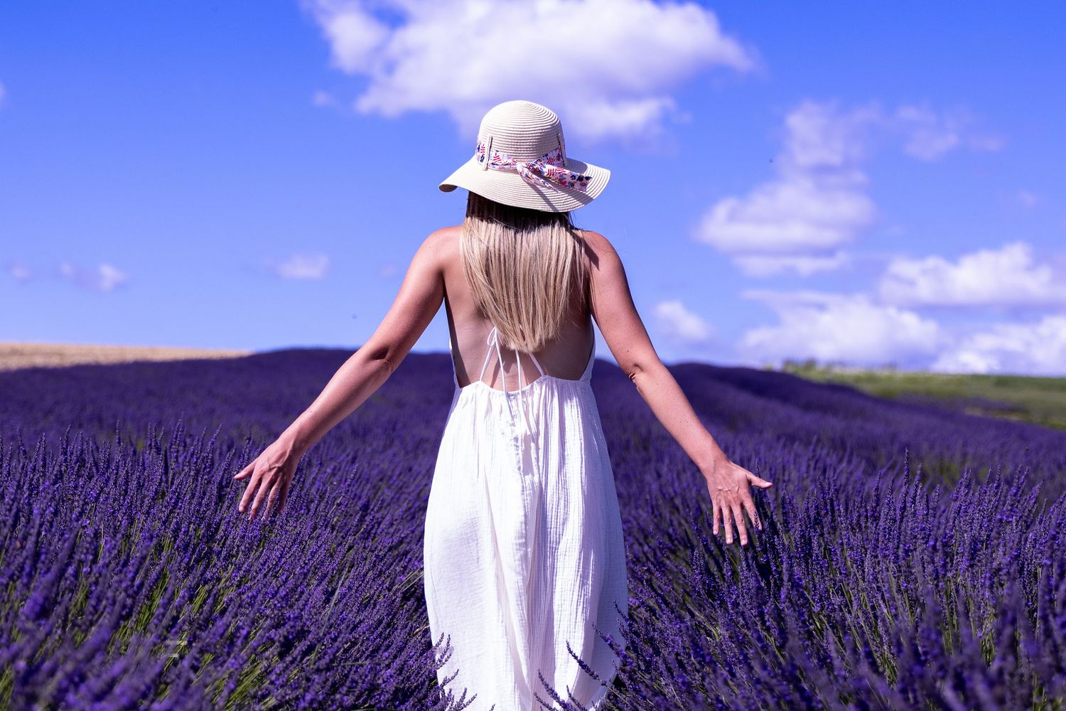 Woman in white dress and hat standing in vibrant purple lavender field under blue sky with clouds.
