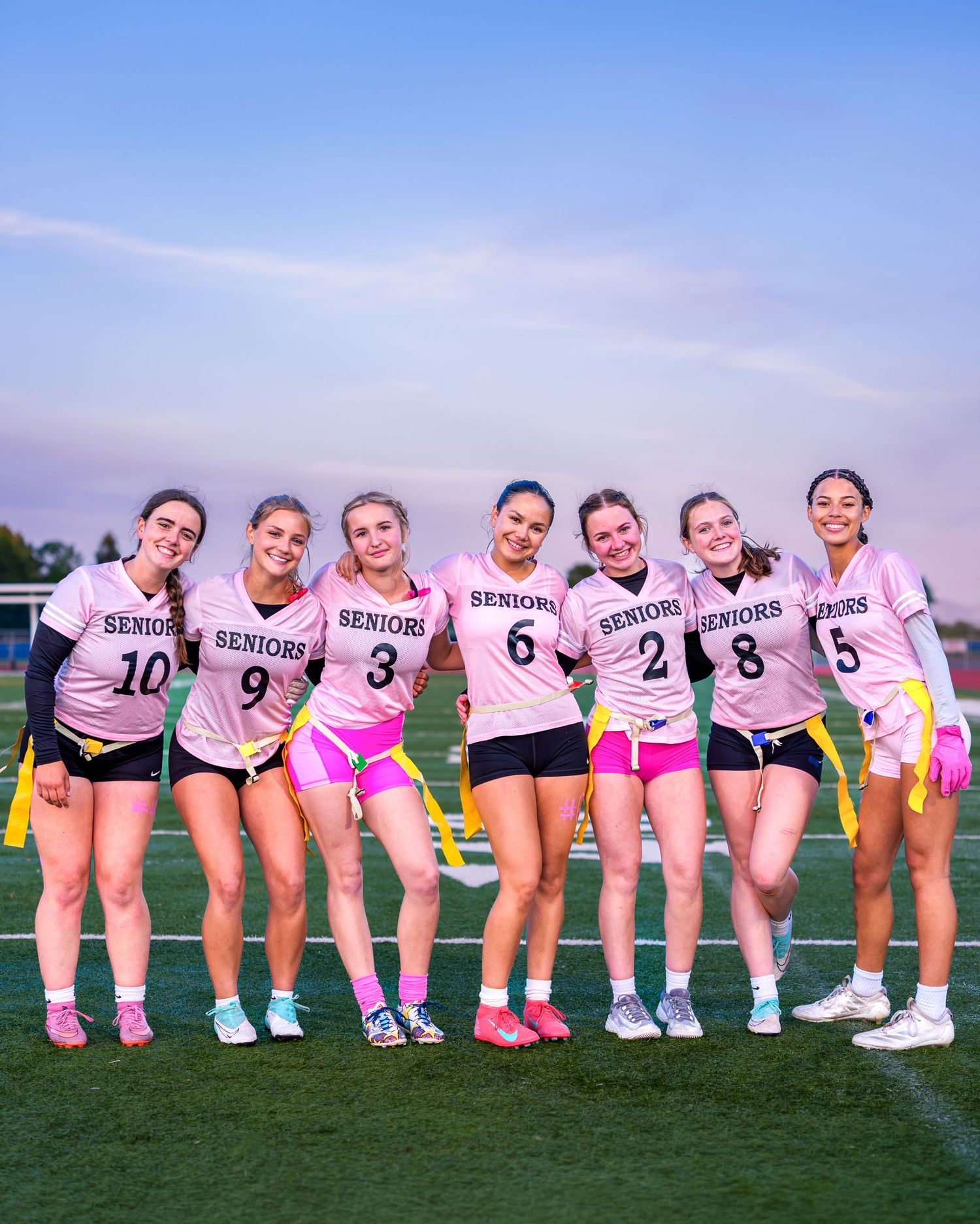 Seven senior female flag football players pose together on a field wearing matching pink jerseys and black shorts.