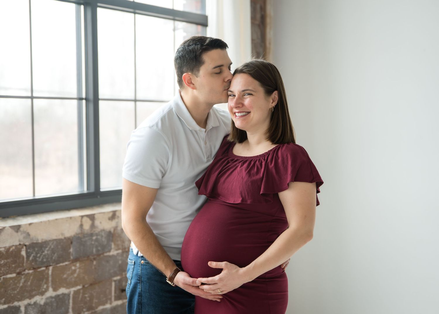 Pregnant woman in burgundy dress cradling her baby bump, smiling down at her belly against a light background.