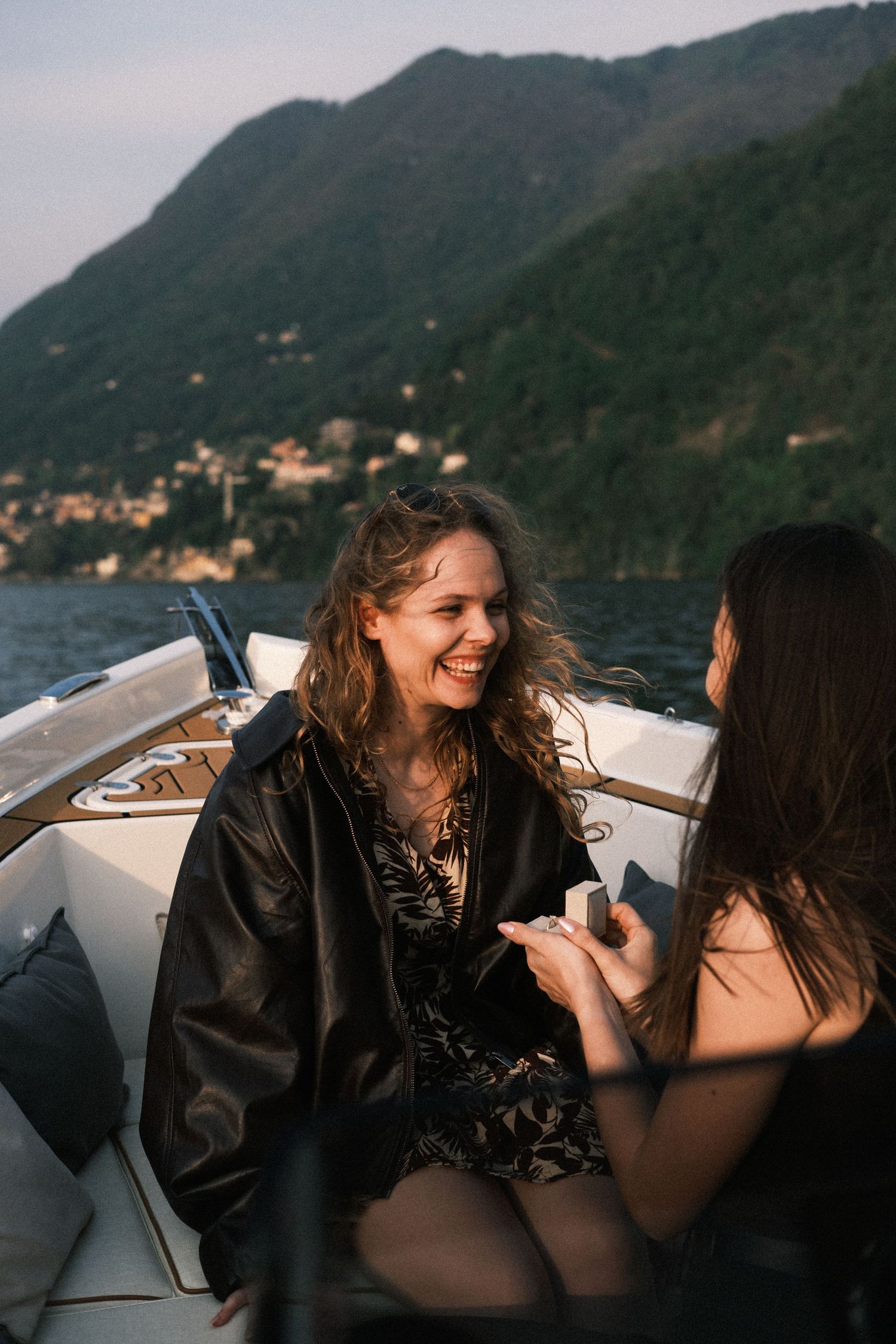 Two women laughing on a luxury boat with scenic mountain lake views in the background at dusk.