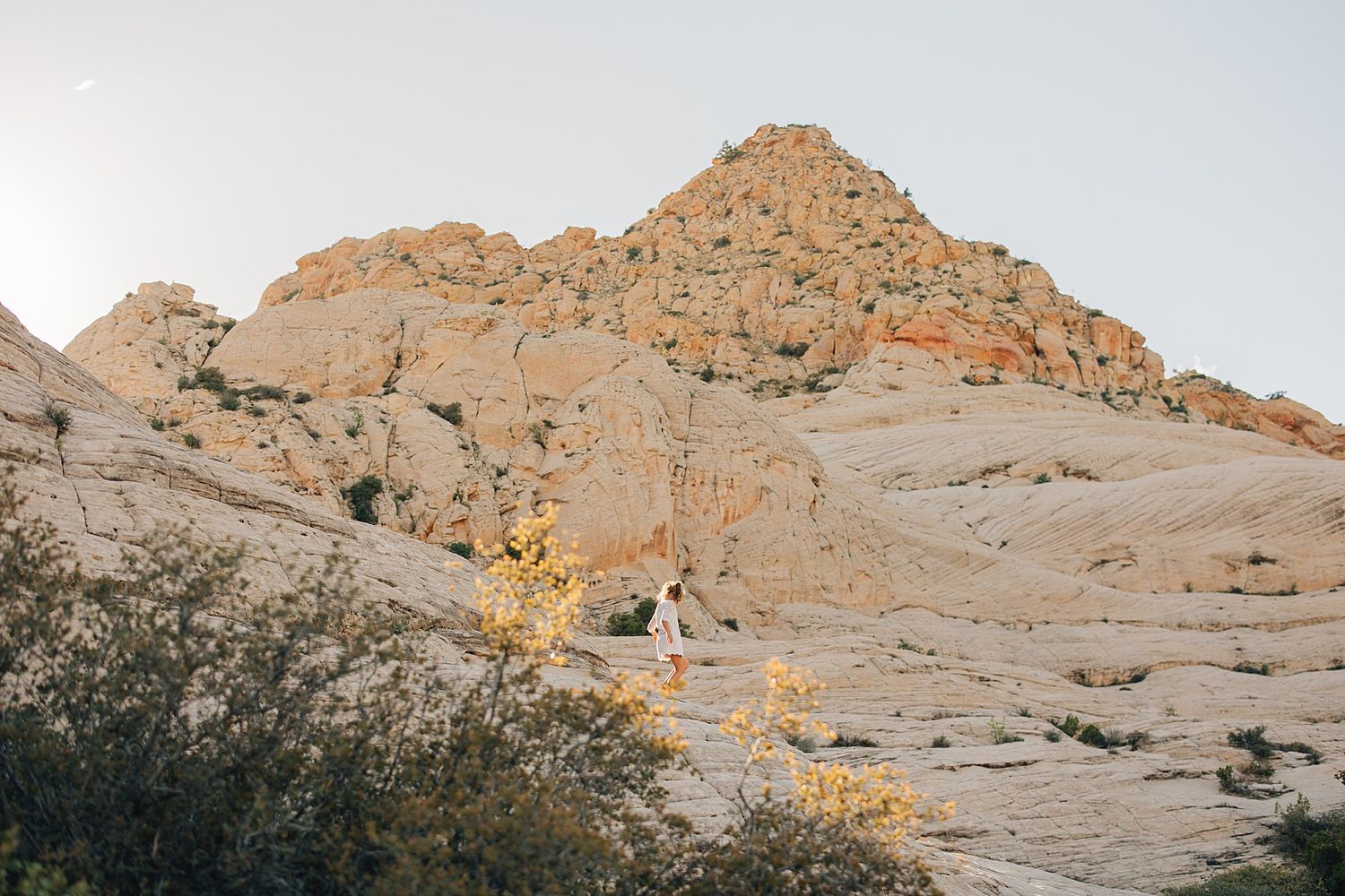 A lone hiker walks along rocky desert terrain beneath a golden sandstone mountain in warm sunlight.
