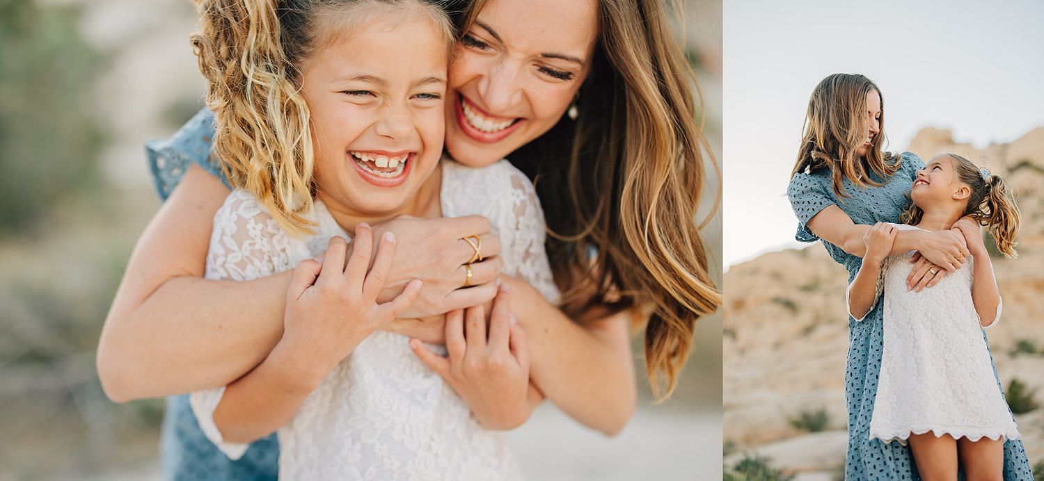 A joyful mother hugs her laughing young daughter outdoors in a sunny, arid landscape, both wearing light dresses.