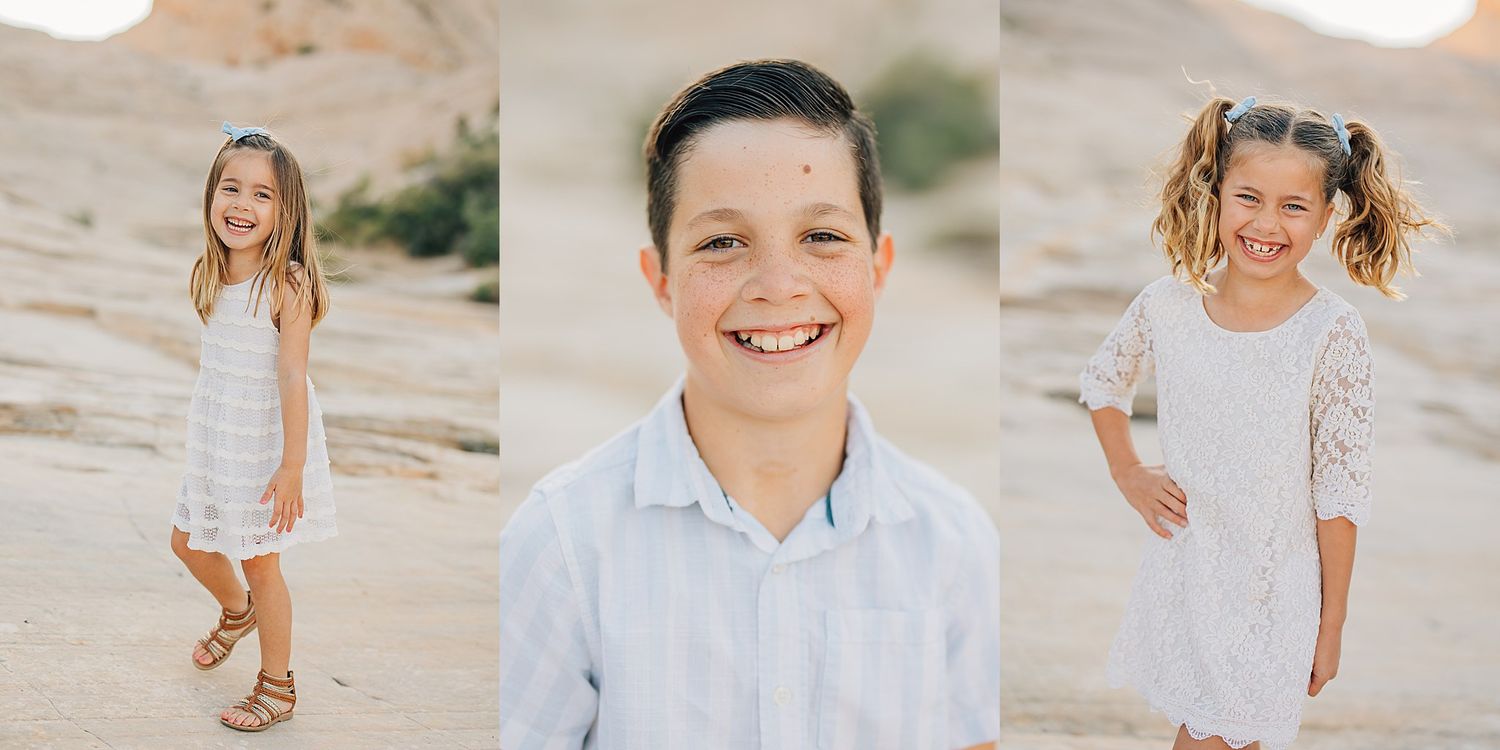 Three children smiling outdoors in desert setting, two girls in white lace dresses and a boy in light blue shirt.