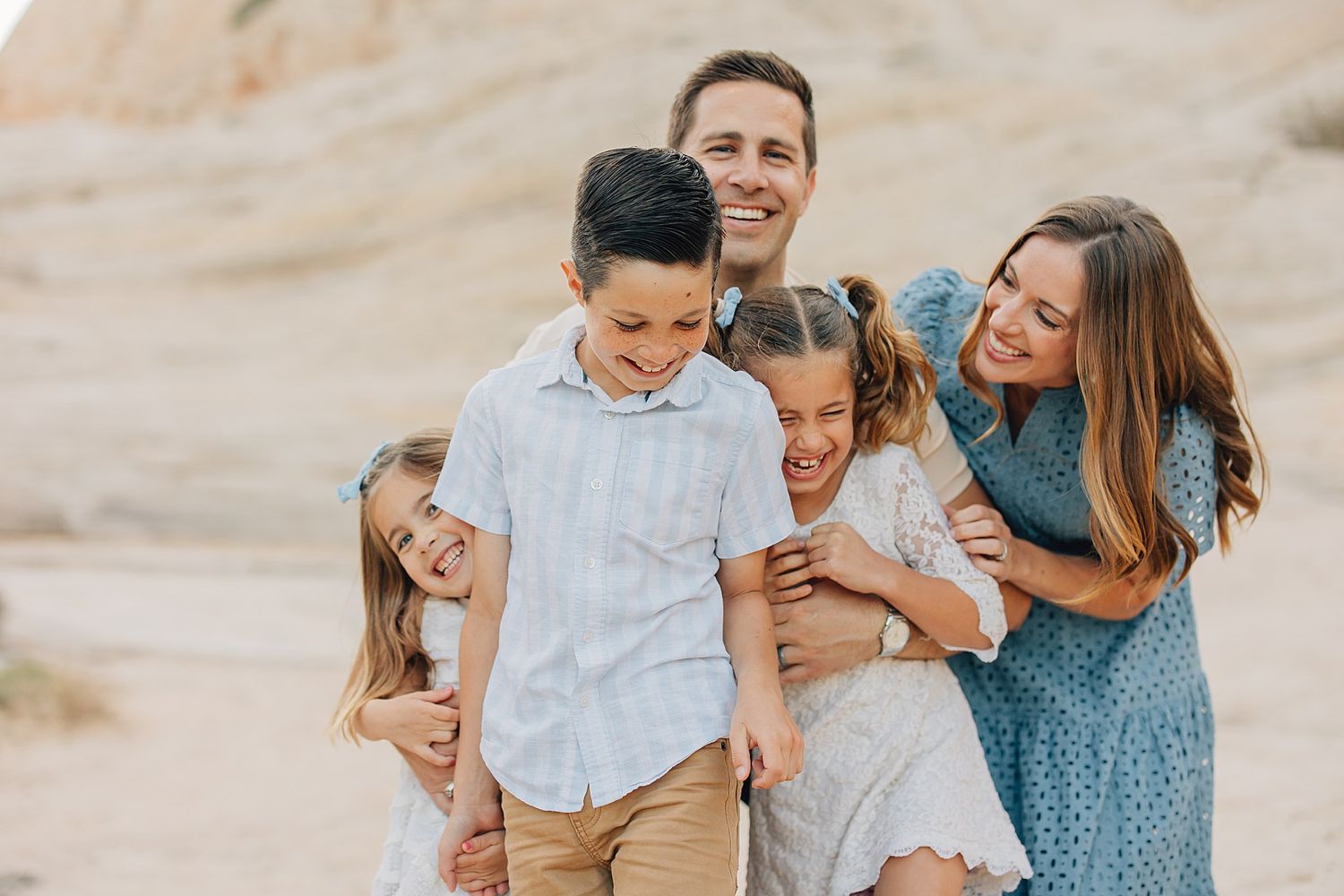 Happy family of five laughing together outdoors in a sandy desert landscape during a professional photo session.
