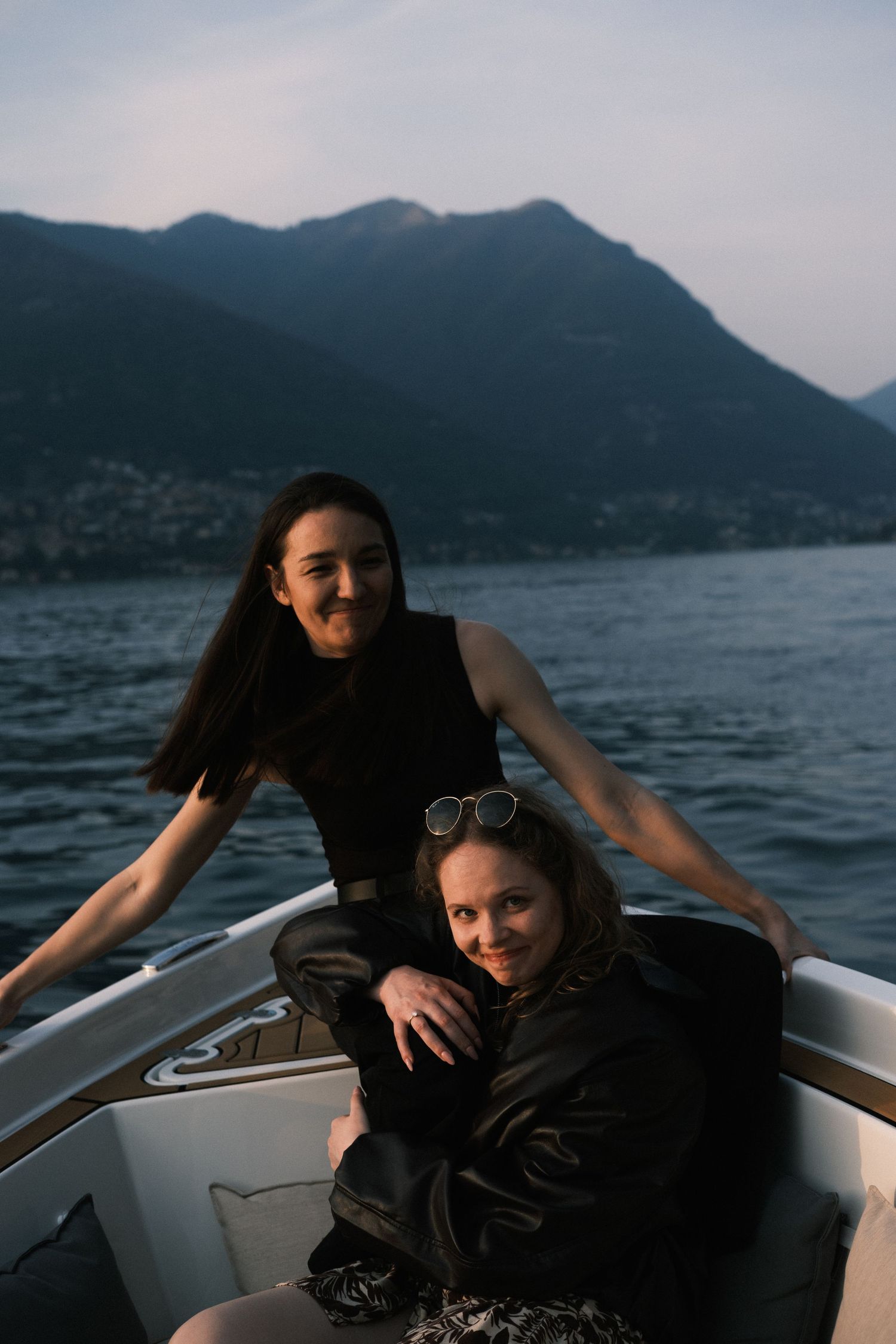 Two women posing on a white motorboat with scenic mountain lake views in the background at dusk.
