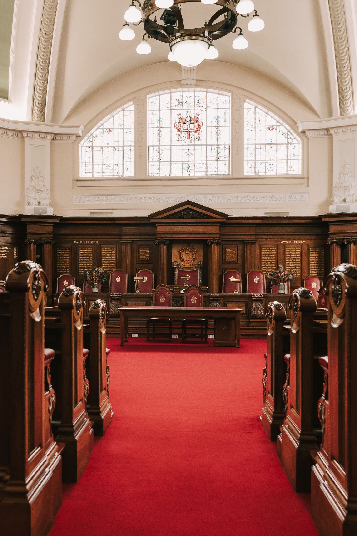 Grand courtroom interior with red carpet, wooden benches, judges' bench, and stained glass window under vaulted ceiling.
