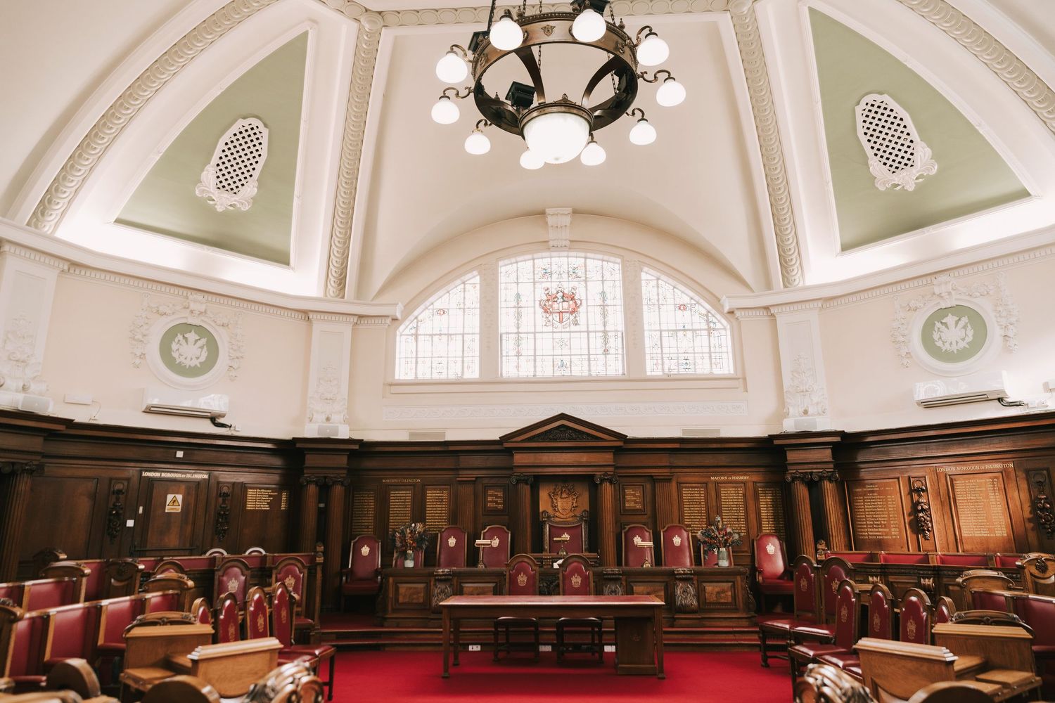 Grand courtroom interior with red carpet, wooden paneling, ornate chandelier, and arched stained glass windows.