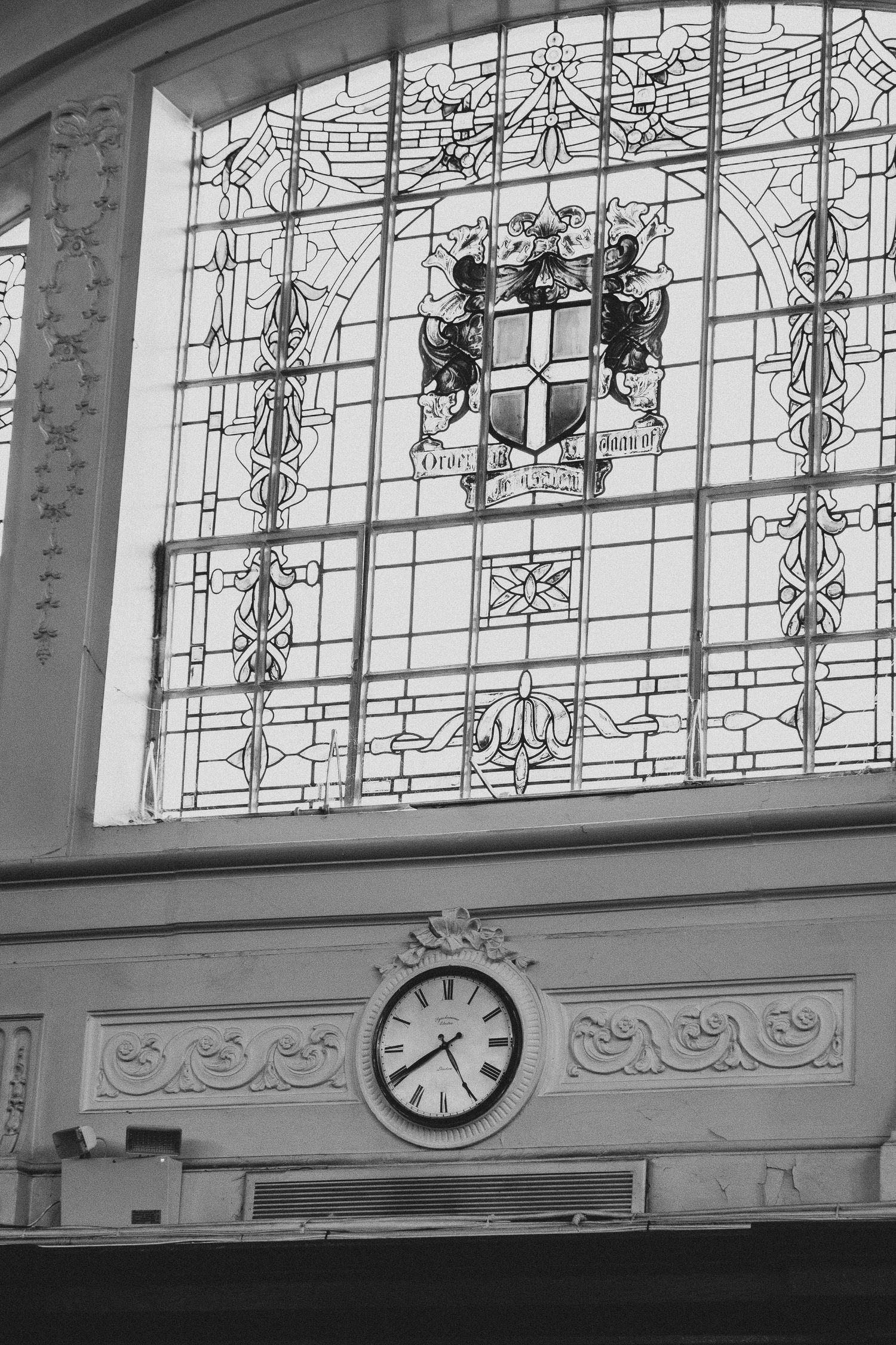 Black and white photo of ornate stained glass window with coat of arms above decorative wall clock in historic building.