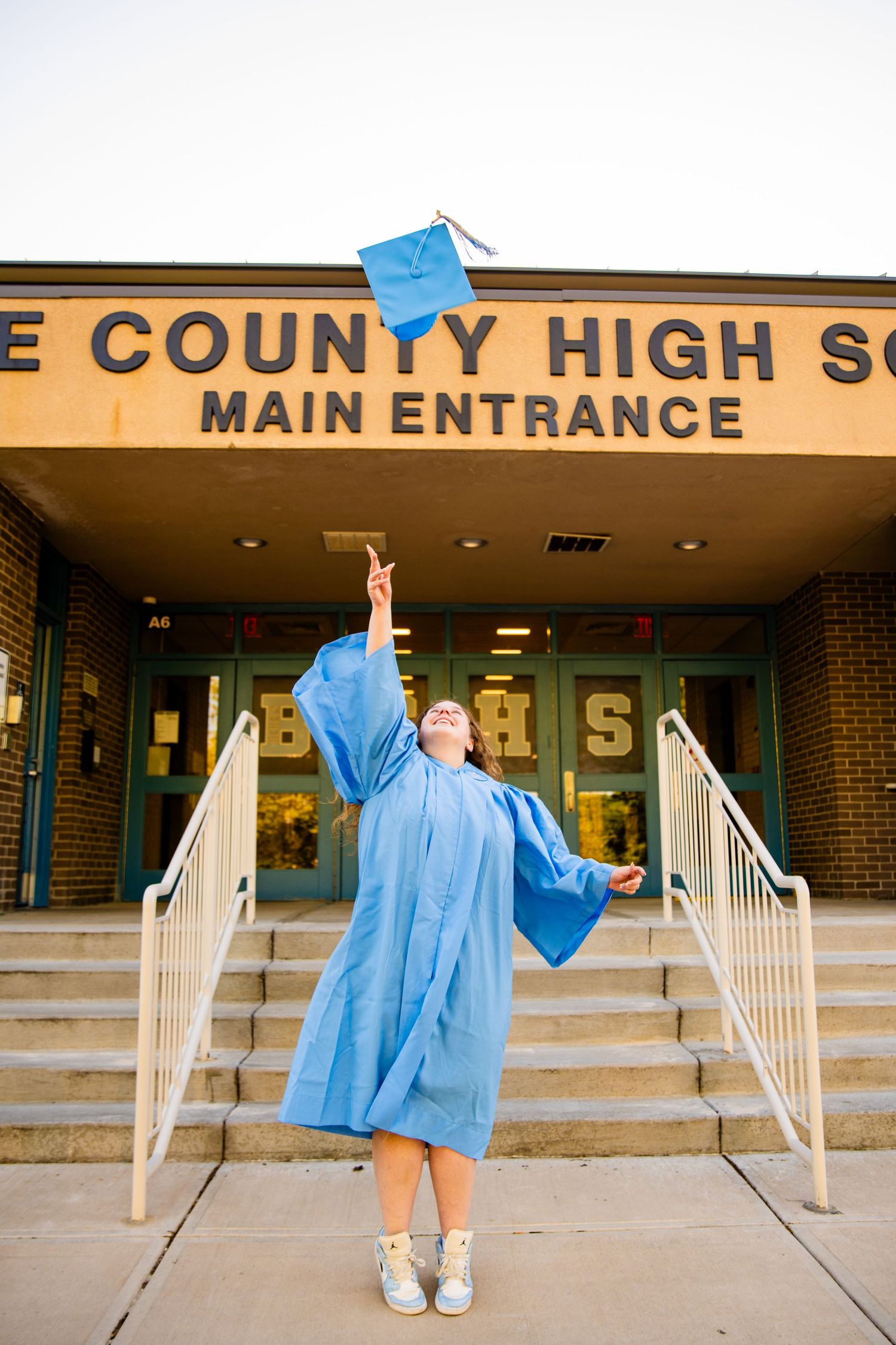 Graduate in blue cap and gown tosses mortarboard in front of County High School main entrance.