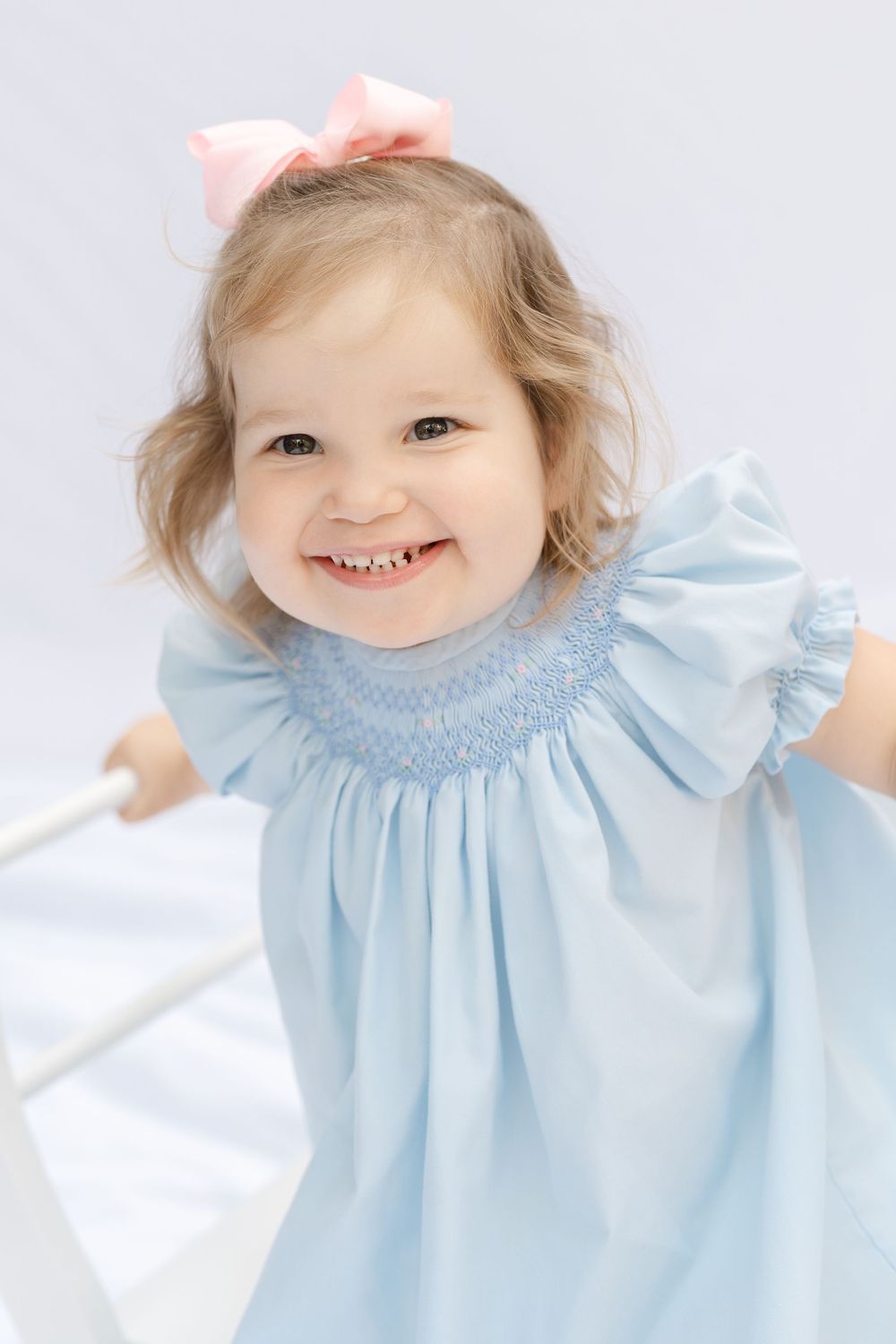 Smiling toddler girl in blue smocked dress with pink bow hair accessory.