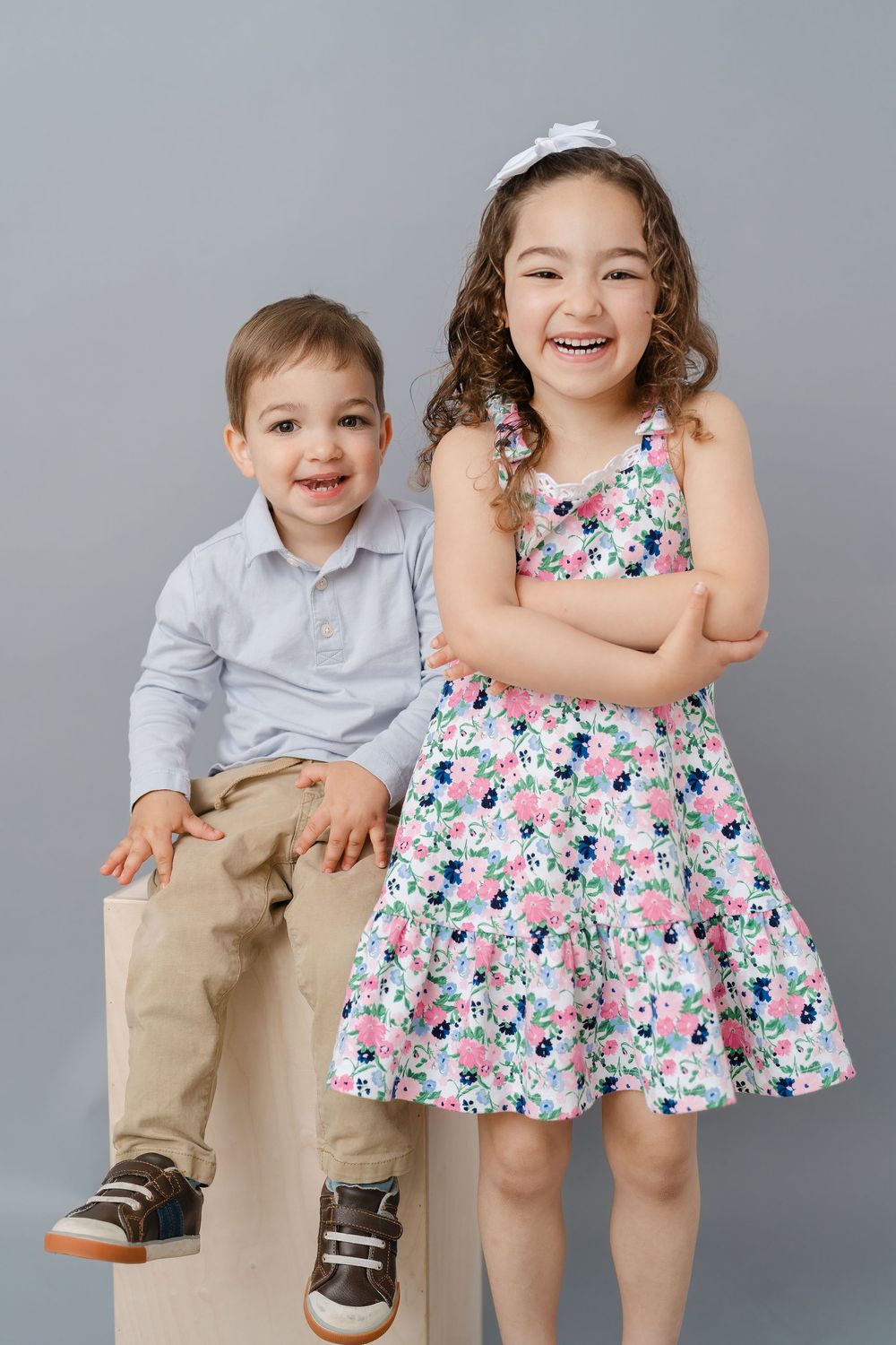Two smiling children, a boy in a polo shirt and a girl in a floral dress, pose together for a studio portrait.