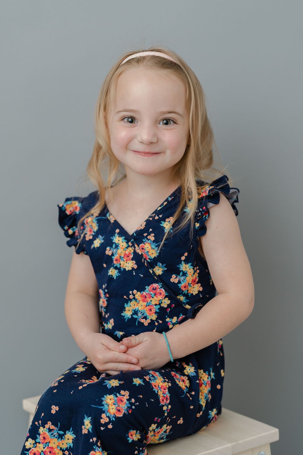 Young girl in navy floral dress sitting and smiling, with blonde hair and headband, against gray background.