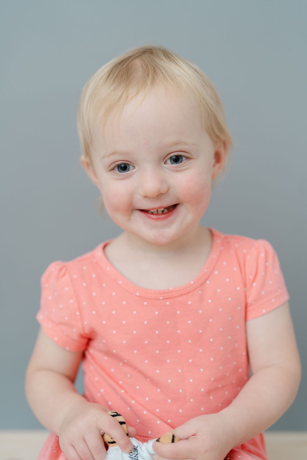 Smiling toddler girl with blonde hair wearing a pink polka dot dress, portrait photo against gray background.