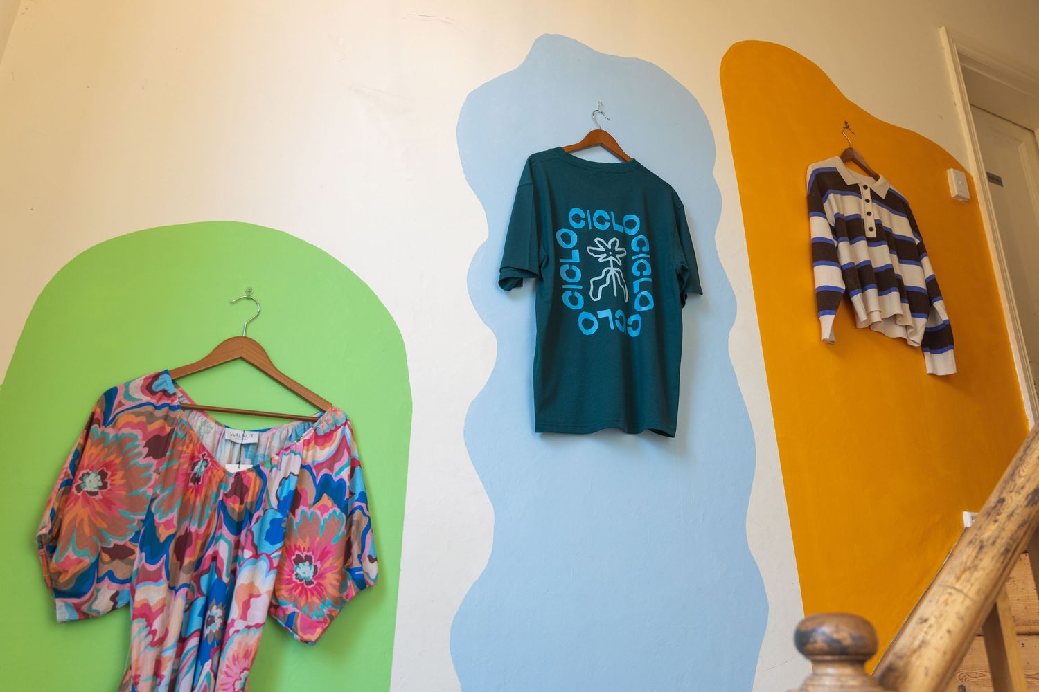 Three colorful shirts displayed on hangers against painted wall art in green, blue, and yellow in a boutique.