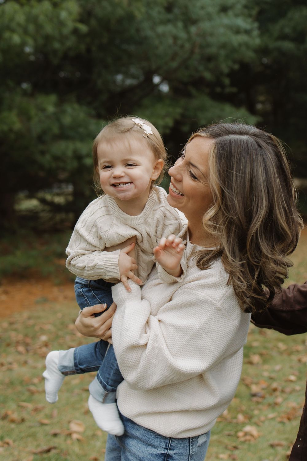Joyful mother in cream sweater holds laughing toddler daughter outdoors among green trees.
