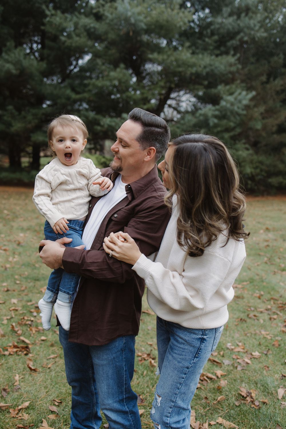 Parents in casual attire laugh with surprised wide-eyed baby boy in autumn park setting.
