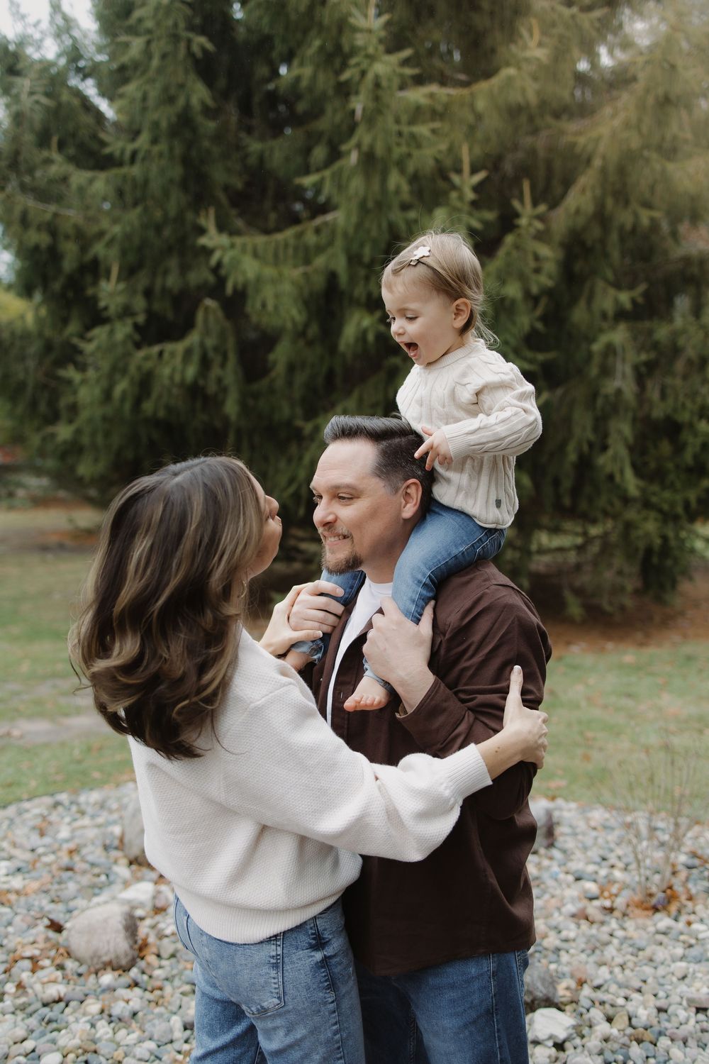 Couple in jeans and casual tops laugh together while father holds excited toddler on shoulders outdoors.