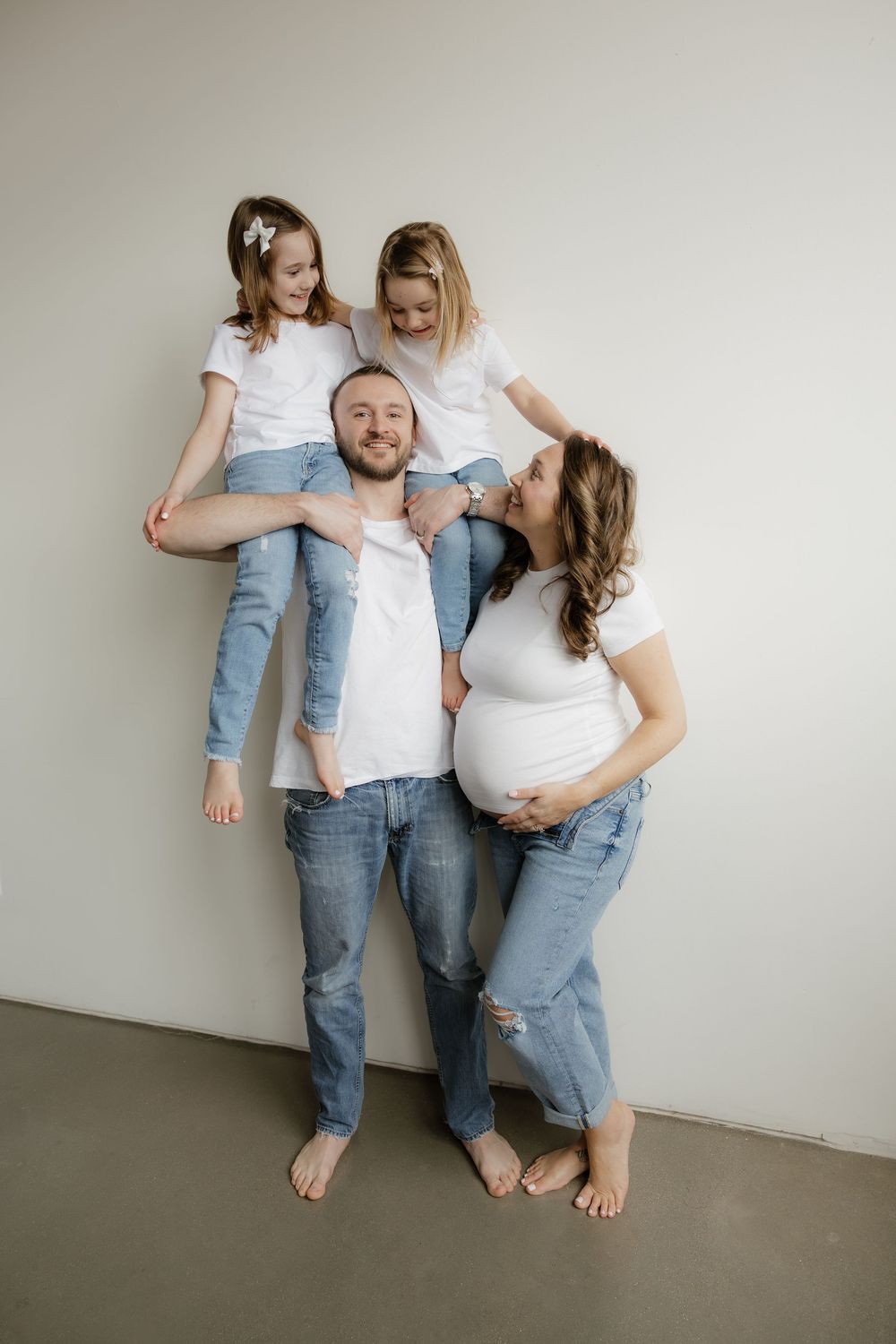 Smiling family in white shirts and jeans pose together, father carrying two daughters on his back.