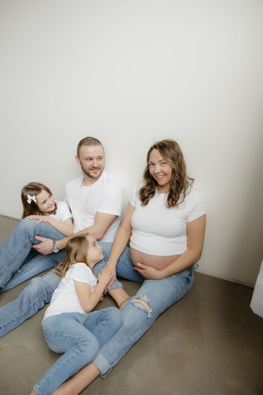 Happy family in matching white shirts and jeans sit on floor with pregnant mother smiling.