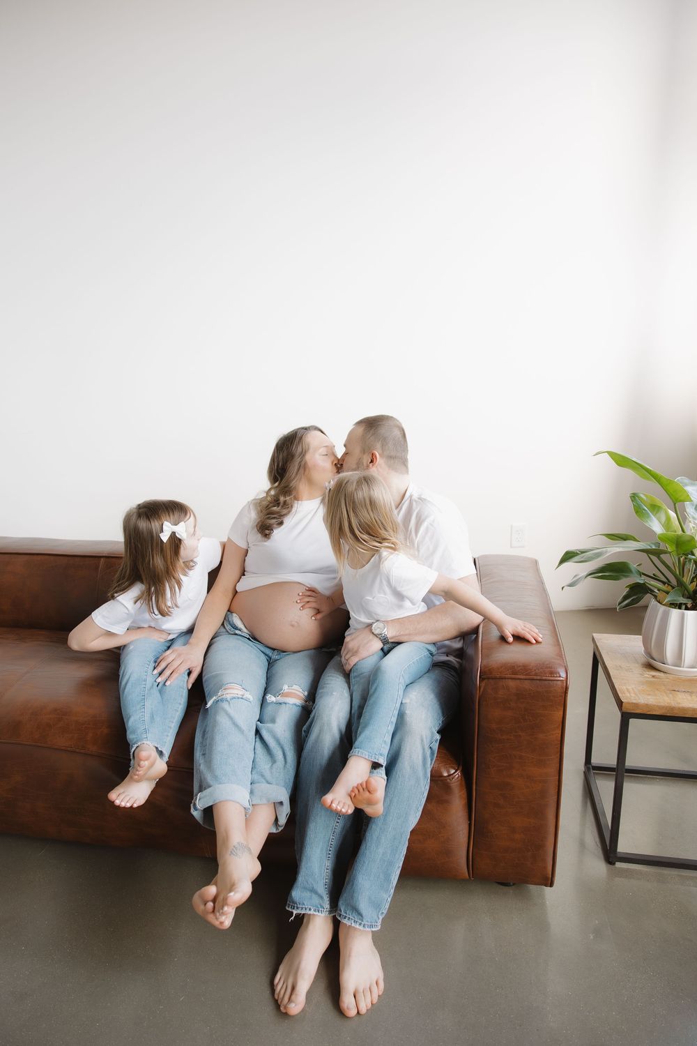 Pregnant woman and husband kiss on leather sofa while two young daughters sit beside them.