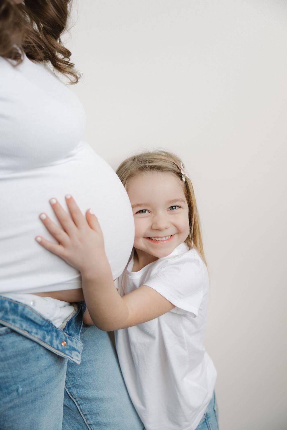 Smiling blonde girl hugs pregnant mothers belly while wearing matching white shirt and jeans.