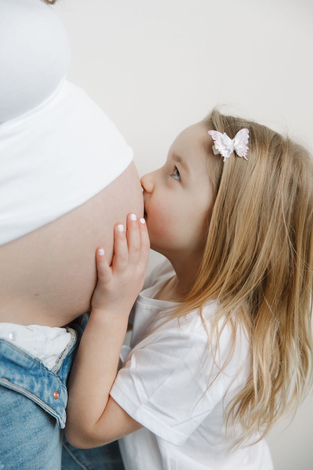 Young blonde girl with butterfly clip kisses pregnant mothers bare belly tenderly.