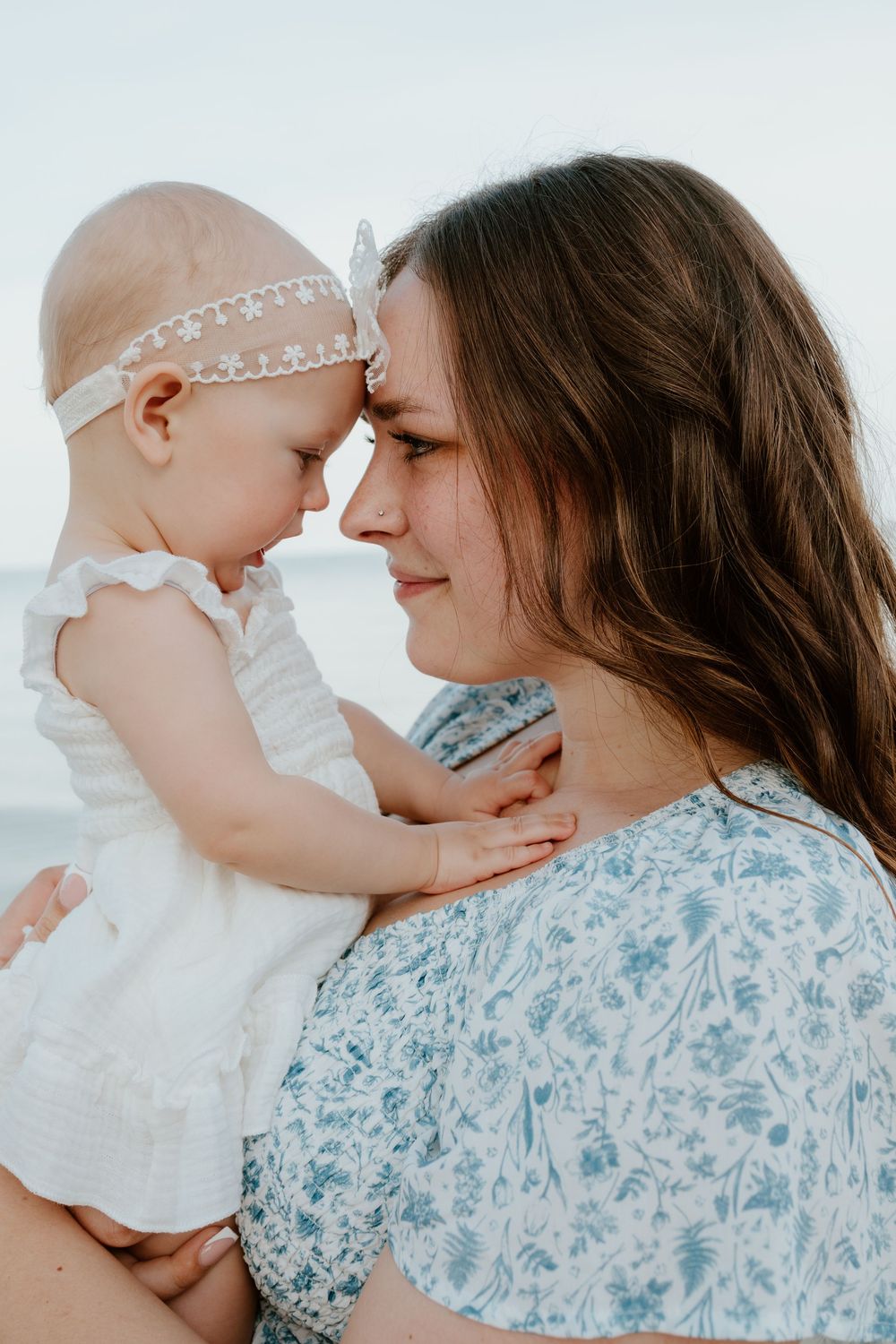 Tender moment of a mother touching foreheads with her baby girl in white dress and headband at beach.