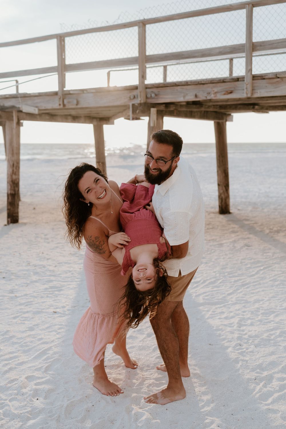 Happy family with young daughter being held upside down laughing on sandy beach near wooden pier.