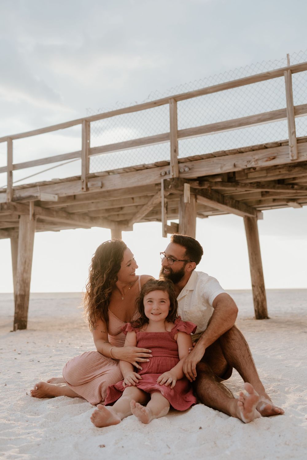 Family of three sitting together on sandy beach near wooden pier, parents gazing lovingly at smiling daughter.