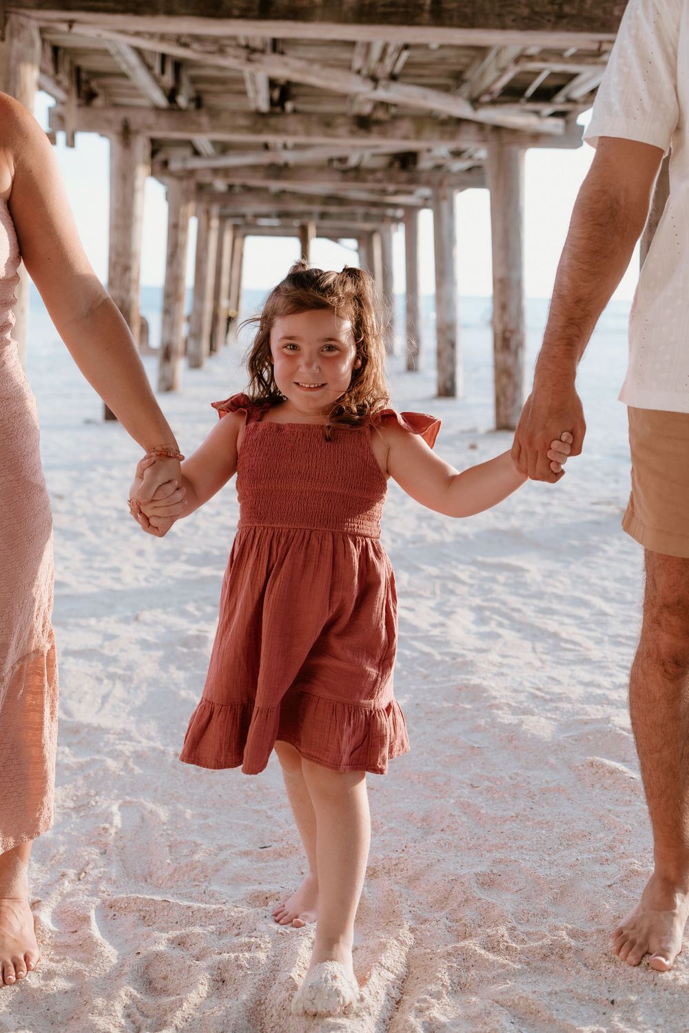 Smiling little girl in red dress holding parents hands while standing under wooden beach pier on white sand.