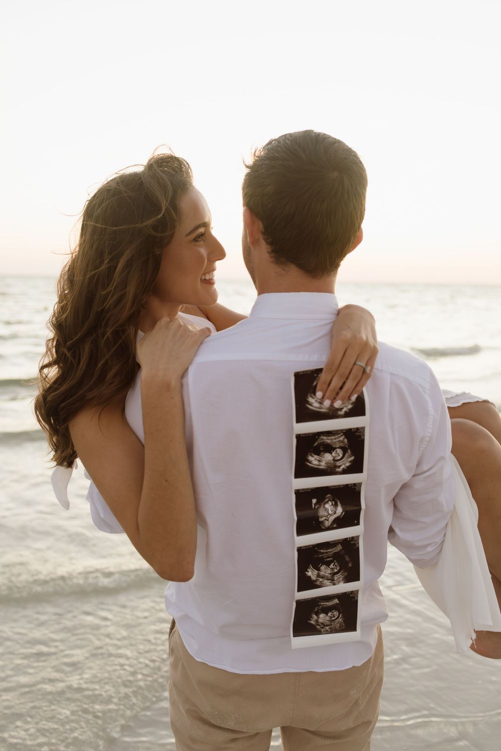 Smiling woman embraces man from behind on beach, showing ultrasound photos on his back at sunset.