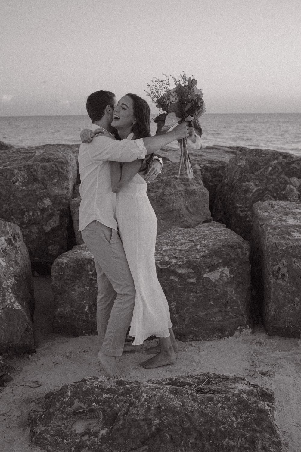 Black and white photo of a joyful couple embracing on rocky beach, woman in white dress holding bouquet laughing.