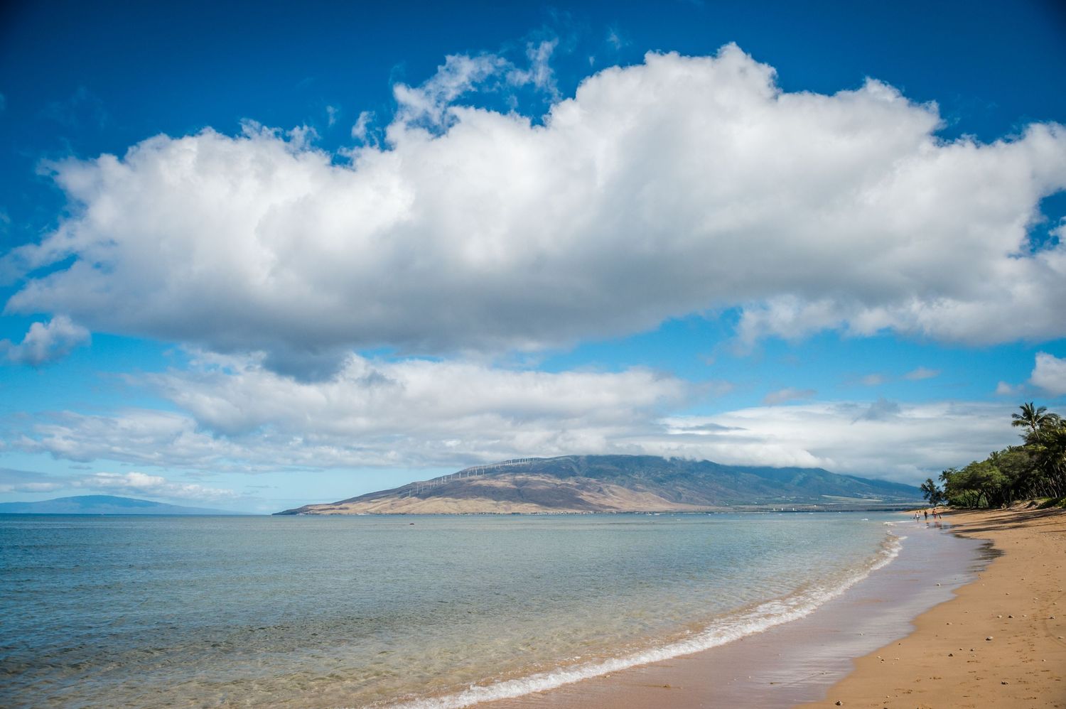 Calm Hawaiian beach with golden sand, turquoise water, lush mountain backdrop, and dramatic white clouds in a blue sky.