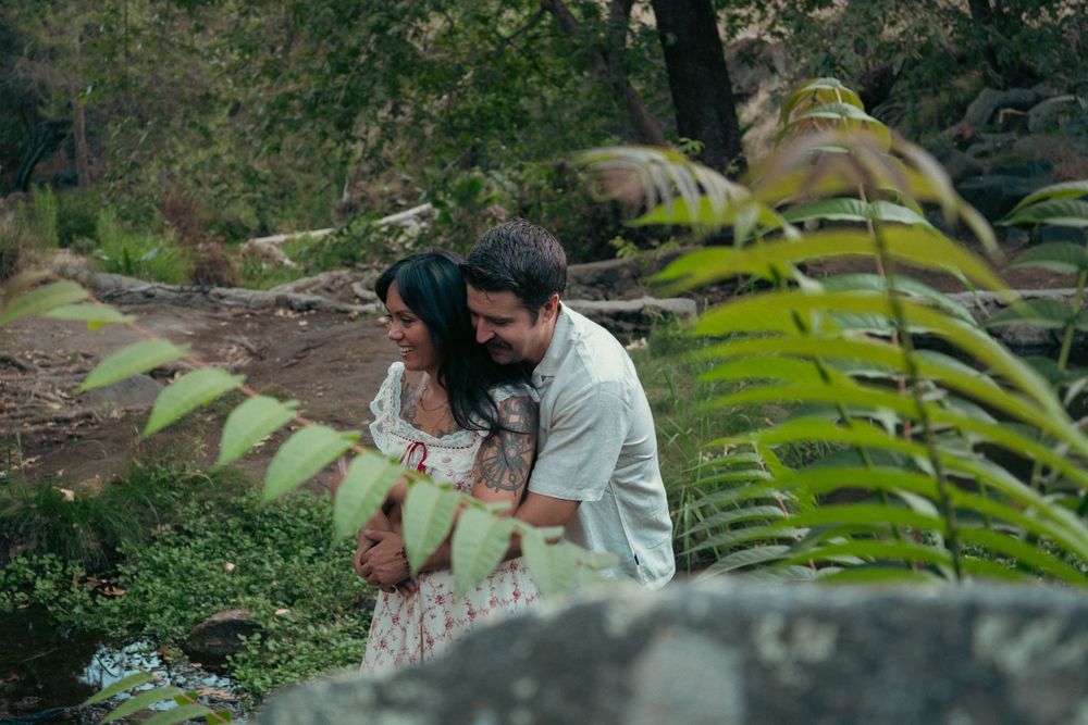 A couple embraces tenderly in a lush green forest setting with ferns and rocks surrounding them. Vista, CA