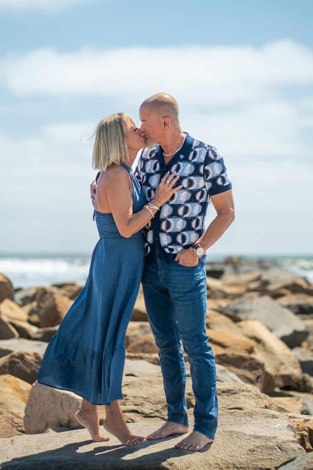 Couple kissing on rocky beach, woman in blue dress and man in patterned shirt, ocean waves in background. Oceanside, CA