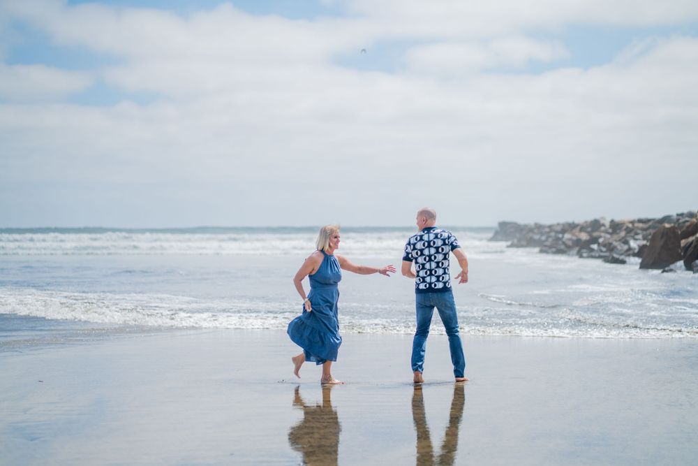 Couple holding hands on a sandy beach with ocean waves, wearing blue outfits on a cloudy day. Oceanside, CA
