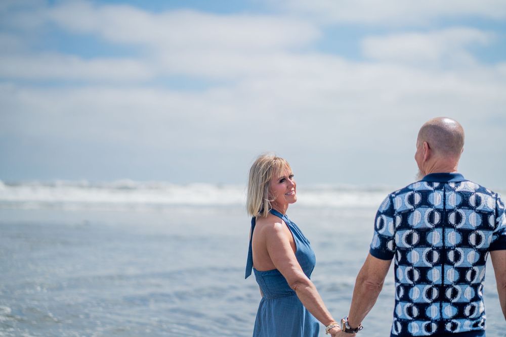 Couple holding hands on a beach, woman in blue dress looking back, man in patterned shirt, ocean waves behind them. Oceanside, CA