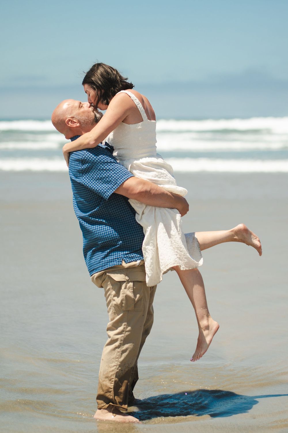 Romantic couple sharing a kiss on the beach, man lifting woman in white dress near ocean waves on sunny day. Oceanside, CA