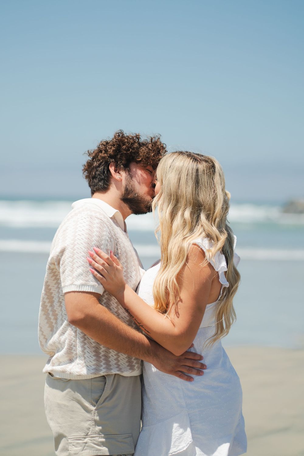 Romantic couple sharing a kiss on a sunny beach, woman in white dress and man in beige shirt embracing by the ocean. Oceanside, CA