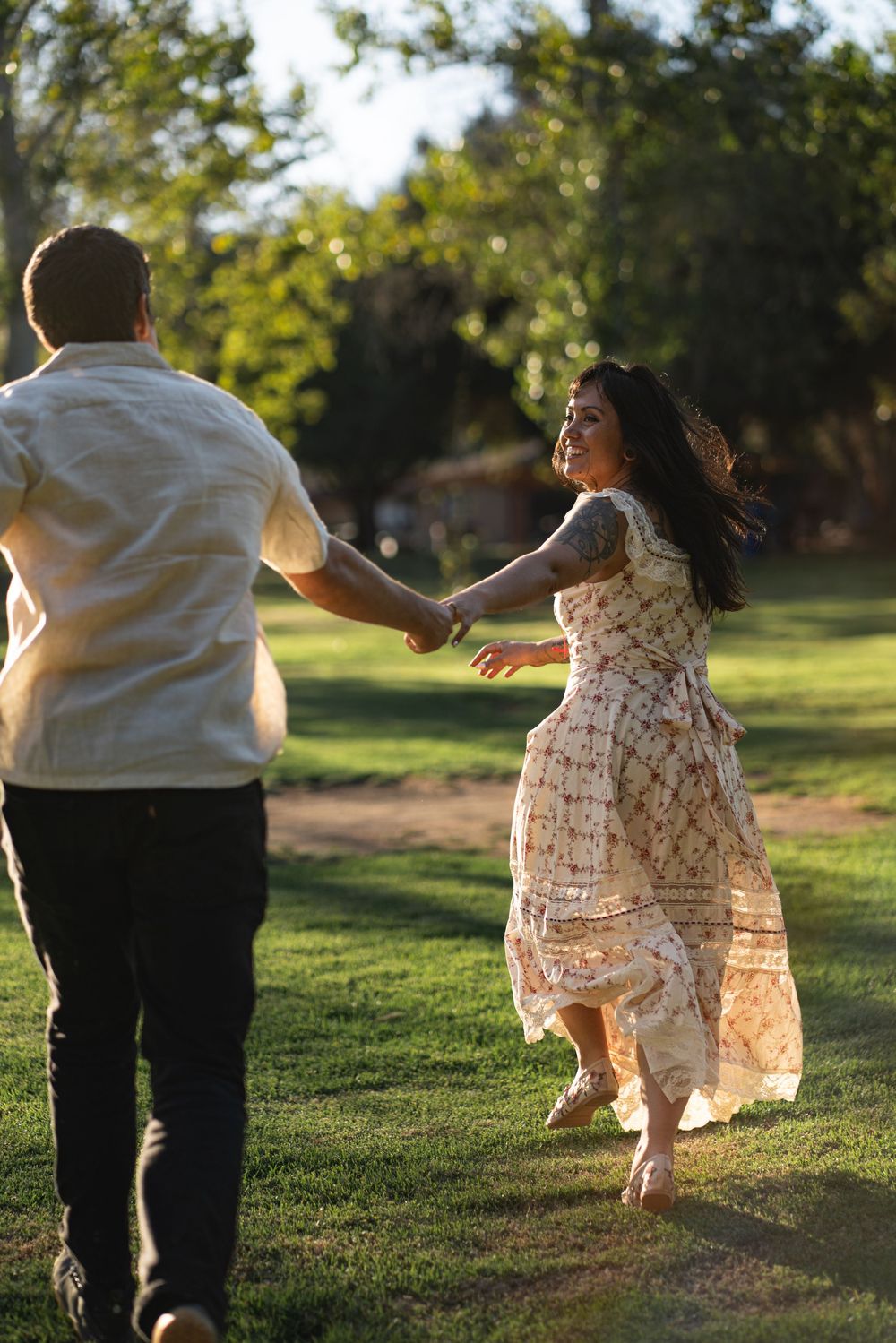A joyful couple holds hands and runs through a sunlit green park, the woman in a floral dress laughing as she looks back. Vista, CA