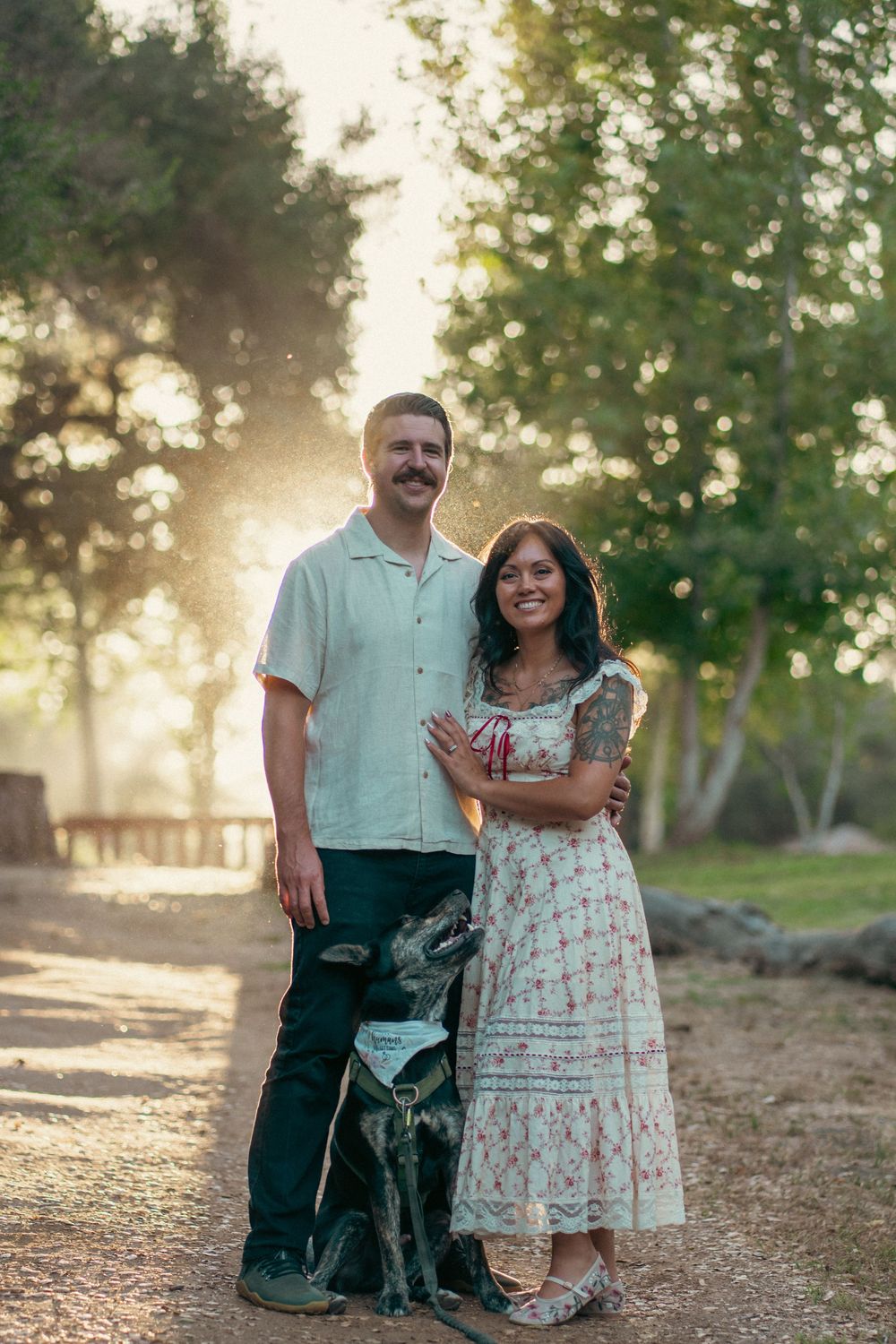 Couple smiling outdoors with their black dog at golden hour, woman in floral dress, man in light shirt, trees in background. Vista, CA